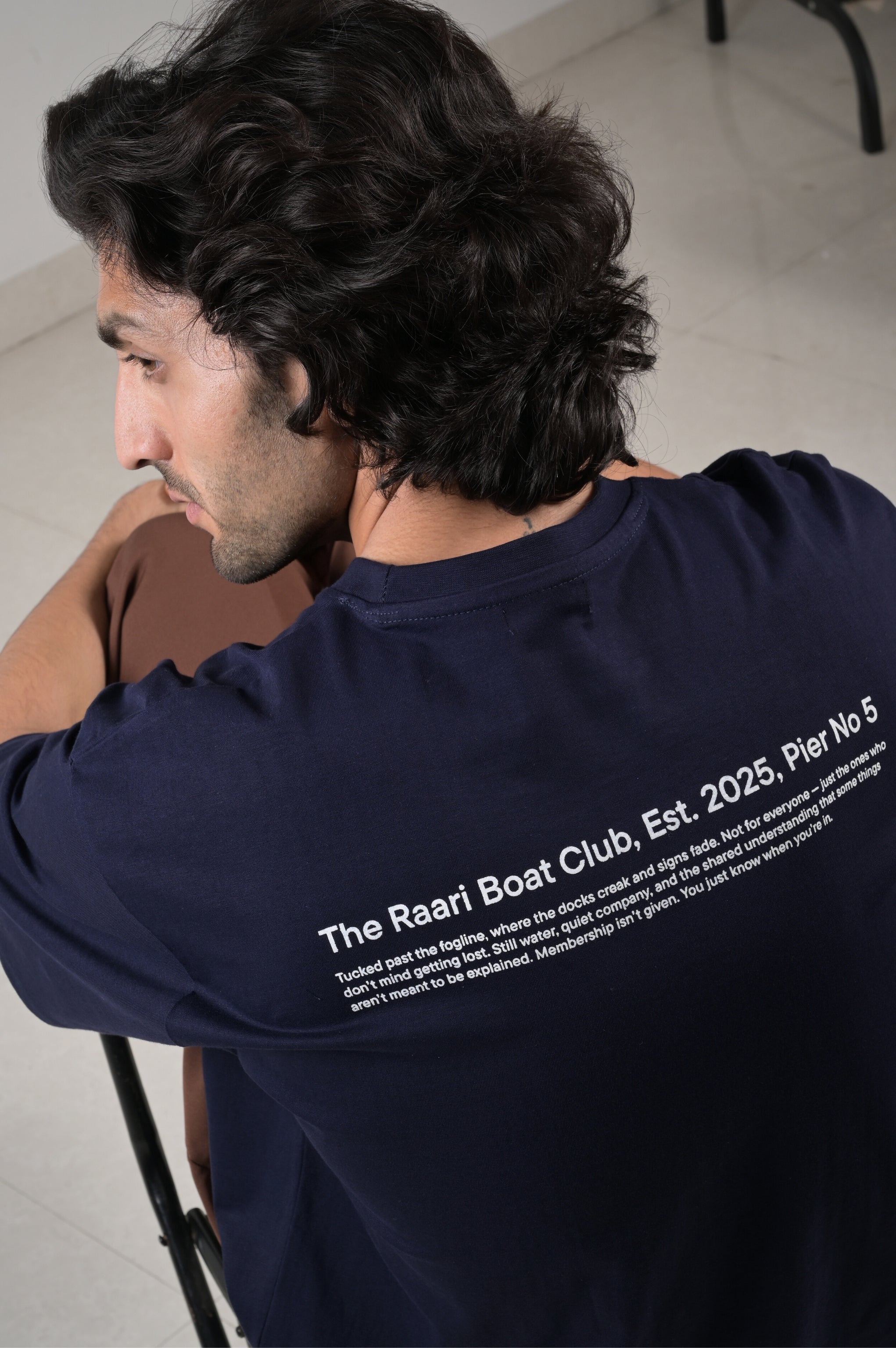 Man wearing a navy blue t-shirt with text on the back, sitting indoors.